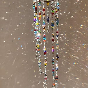 May include: A hanging mobile featuring a circular silver-toned metal ring from which multiple strands of multicolored faceted glass beads are suspended. Each strand terminates in a clear, teardrop-shaped crystal pendant, creating a light-catching display.