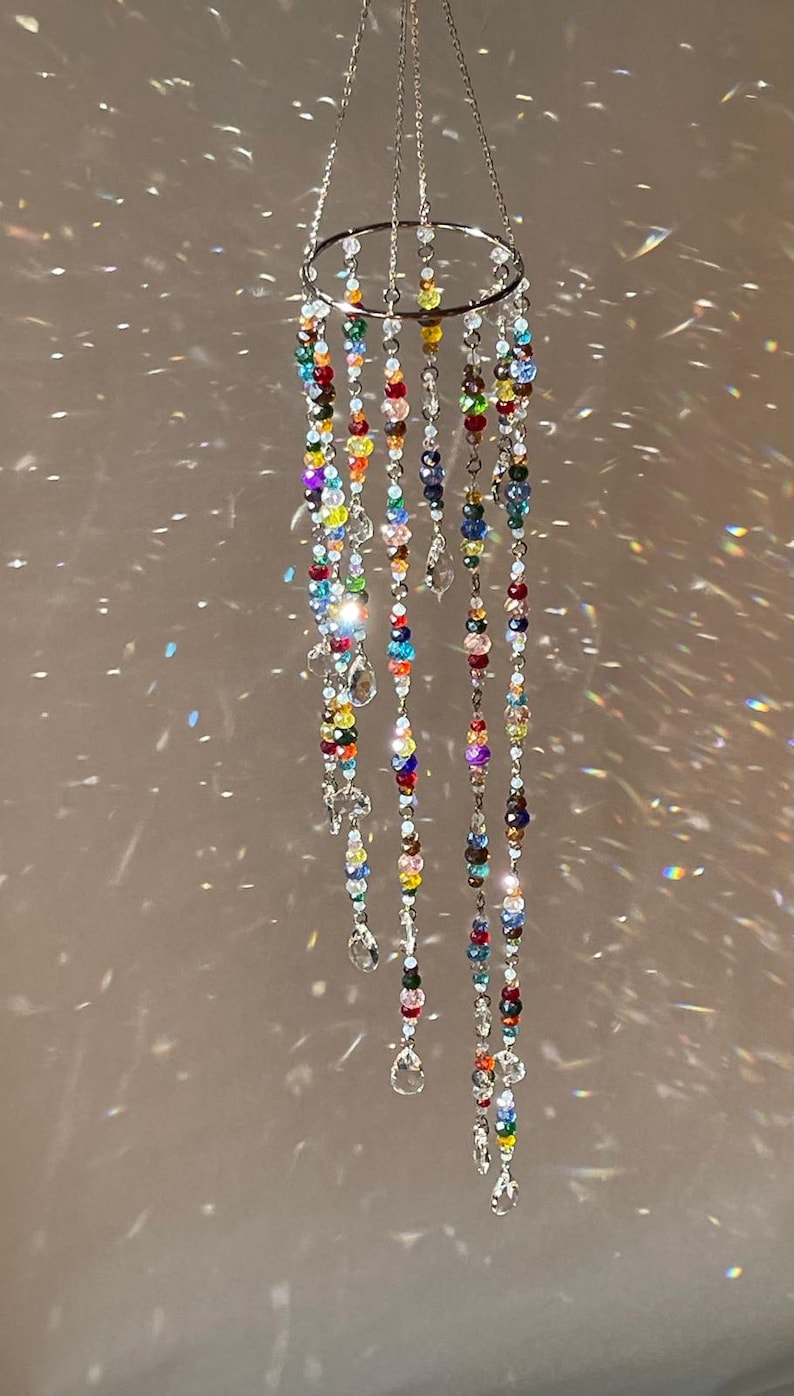 May include: A hanging mobile featuring a circular silver-toned metal ring from which multiple strands of multicolored faceted glass beads are suspended. Each strand terminates in a clear, teardrop-shaped crystal pendant, creating a light-catching display.