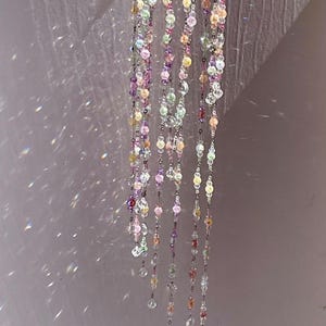 May include: A hanging mobile featuring a circular silver-toned metal frame with multiple dangling strands of multicolored translucent beads in shades of pink, purple, yellow, and clear, creating light reflections on the surrounding surface.