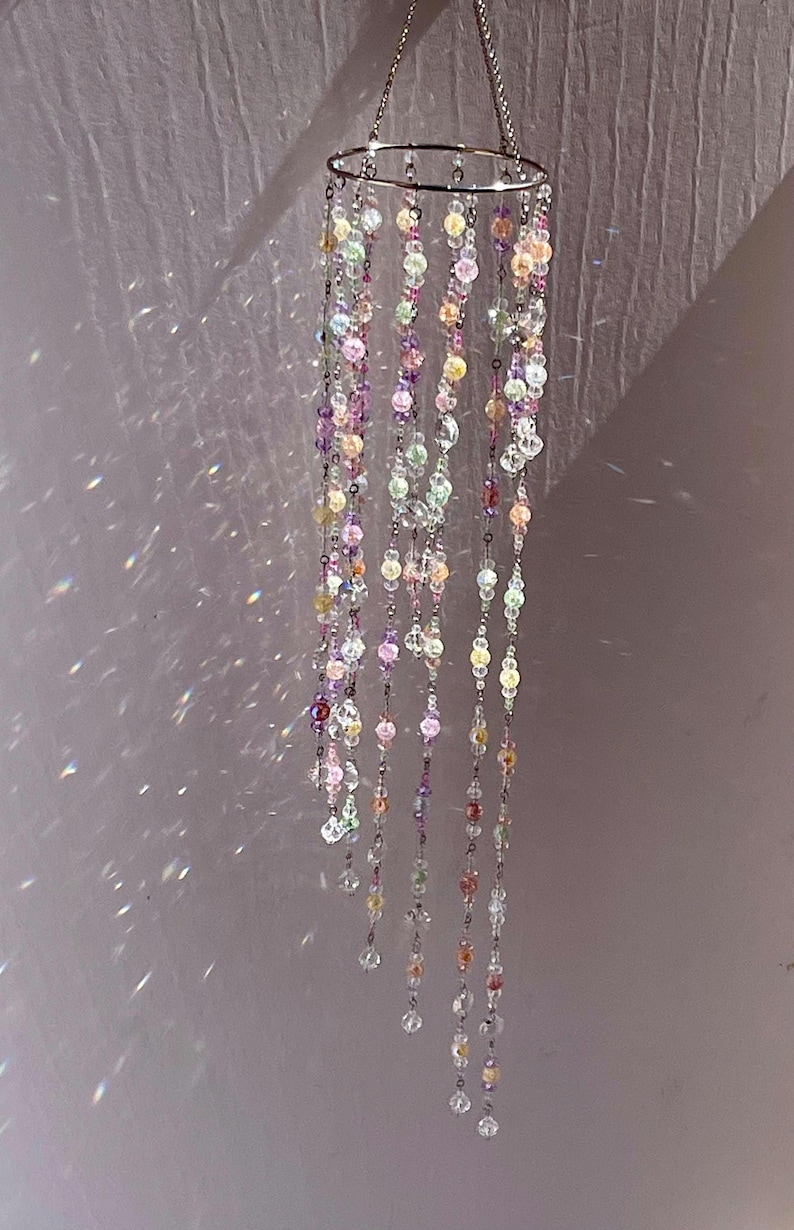 May include: A hanging mobile featuring a circular silver-toned metal frame with multiple dangling strands of multicolored translucent beads in shades of pink, purple, yellow, and clear, creating light reflections on the surrounding surface.