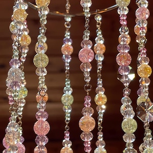 May include: A hanging mobile featuring multiple vertical strands of beads suspended from a circular metal ring. The strands consist of clear, faceted crystal beads interspersed with round, crackled glass beads in shades of pink, yellow, and green.