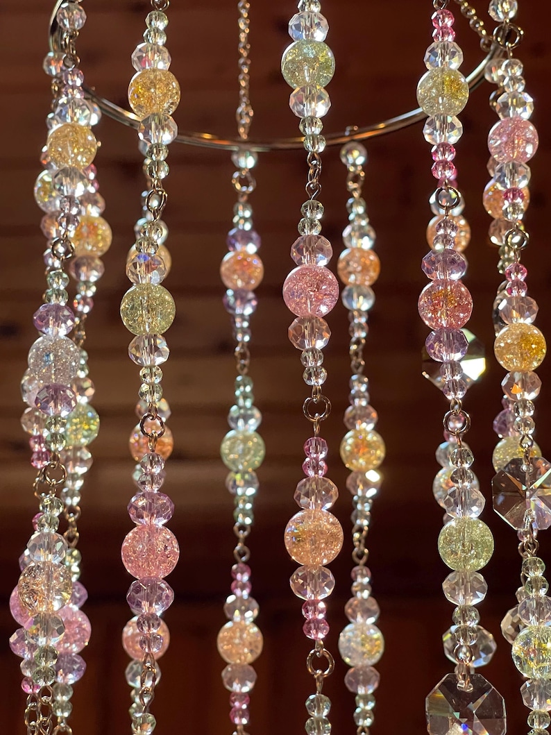 May include: A hanging mobile featuring multiple vertical strands of beads suspended from a circular metal ring. The strands consist of clear, faceted crystal beads interspersed with round, crackled glass beads in shades of pink, yellow, and green.
