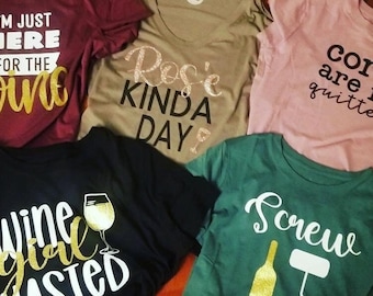 May include: A collection of five t-shirts with wine-themed graphics and text. The shirts are in various colors, including burgundy, olive green, black, pink, and green. Text includes "I'm just here for the wine", "Rosé Kinda Day", "Wine Girl Wasted", "Cork are quitters", and "Screw".