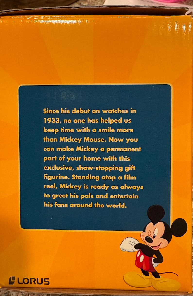May include: A box with a blue panel featuring text about Mickey Mouse and a cartoon image of Mickey. The box is orange and has the Lorus logo at the bottom. The text mentions Mickey's debut on watches in 1933.
