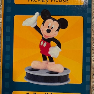 May include: A collectible Mickey Mouse figure on a film reel base, with a yellow and blue background. The figure is wearing red shorts, white gloves, and large pink shoes. Text on the box reads "The One and Only Mickey Mouse" and "Collectible Gift."