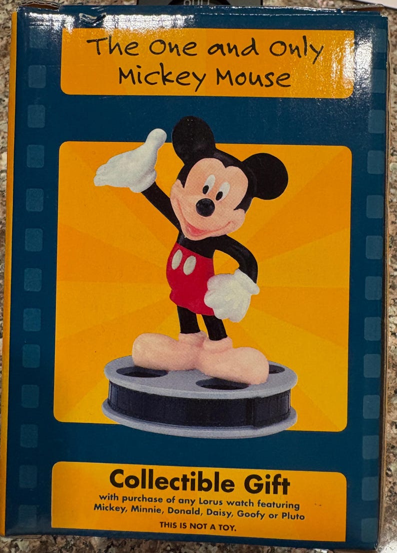 May include: A collectible Mickey Mouse figure on a film reel base, with a yellow and blue background. The figure is wearing red shorts, white gloves, and large pink shoes. Text on the box reads "The One and Only Mickey Mouse" and "Collectible Gift."