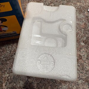 May include: White styrofoam packaging with molded compartments, wrapped in clear plastic. The packaging is designed to hold a product, with circular and rectangular indentations. A portion of a blue and yellow box is visible.