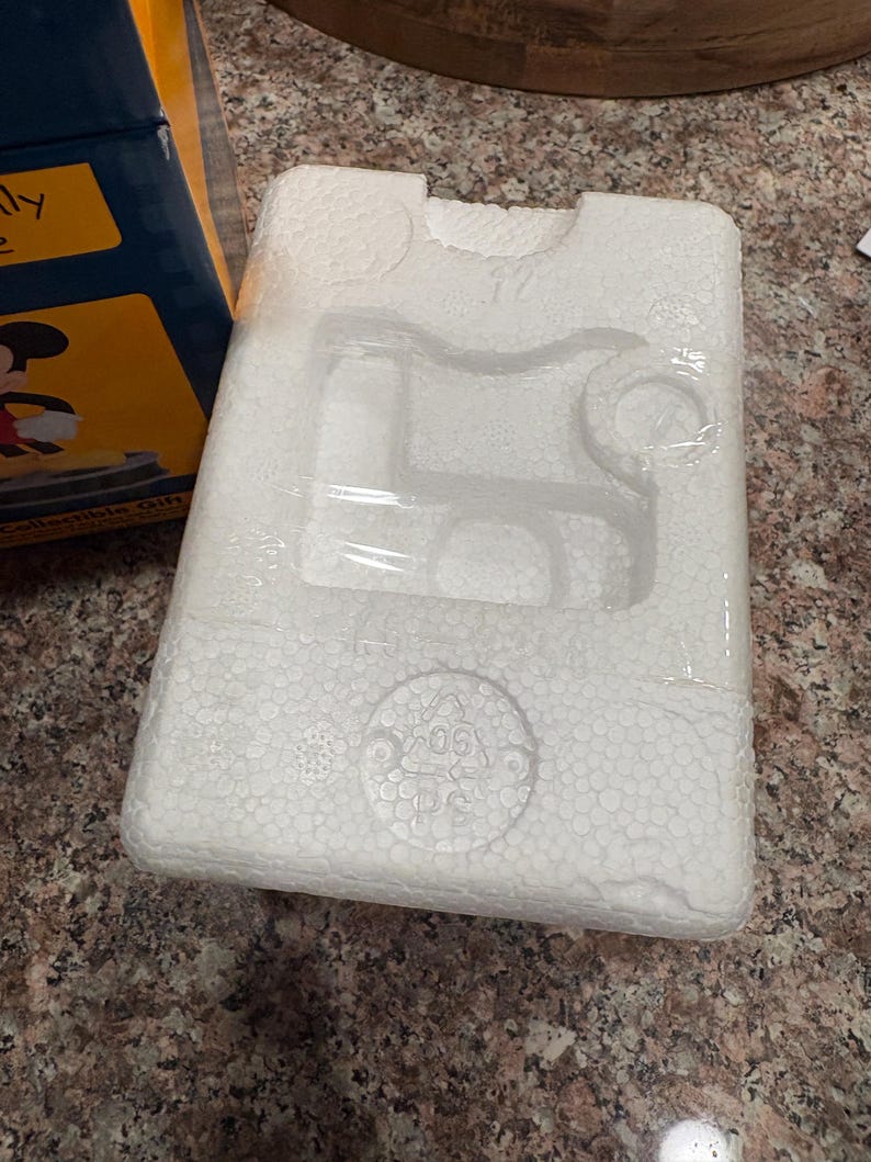 May include: White styrofoam packaging with molded compartments, wrapped in clear plastic. The packaging is designed to hold a product, with circular and rectangular indentations. A portion of a blue and yellow box is visible.