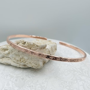 Peut inclure: Un bracelet manchette en métal de couleur or rose avec un design fin et ouvert. La surface extérieure du bracelet est ornée d'un motif gravé répétitif composé de petites fleurs délicates qui s'étendent sur toute la longueur.