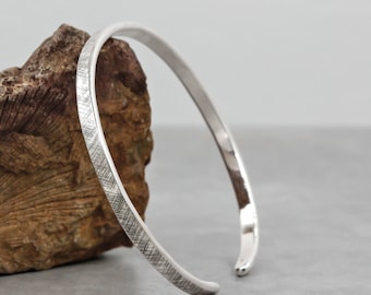 Puede incluir: Un brazalete de plata con un acabado texturizado. El brazalete está abierto y tiene un diseño simple.