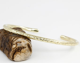 May include: A gold hammered bangle bracelet. The bracelet is made of a thick, textured metal and has a simple, elegant design.