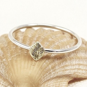 May include: A silver ring with a gold leaf design. The leaf is centered on the ring and has detailed veining. The ring is set against a neutral background, highlighting the jewelry's delicate features. This ring is a nature-inspired accessory.