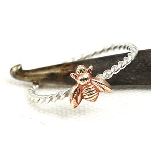 Puede incluir: Un anillo de plata con una banda trenzada y un dije de abeja en oro rosa. La abeja tiene alas y cuerpo detallados, y el anillo se presenta sobre un fondo neutro. Una delicada pieza de joyería.
