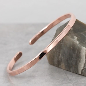May include: A rose gold cuff bracelet with a hammered texture. The open-ended bracelet is displayed on a grey stone, showcasing its polished finish and simple design. This piece of jewellery is a stylish accessory.