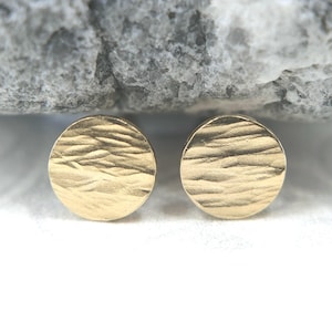May include: A pair of gold-colored stud earrings. Each earring is a small, textured circle. The earrings are set against a neutral background, with a large, gray rock in the background.