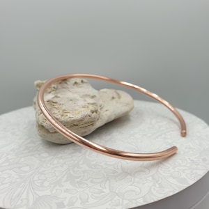 May include: A copper-coloured, open-ended bangle bracelet. The bracelet is round and rests on a light-coloured, textured stone. The background is a light grey. The bracelet is a simple, elegant piece of jewellery.