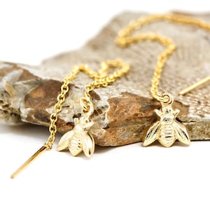 May include: A pair of gold bee charm earrings with a delicate chain. The earrings are hanging from a rock.