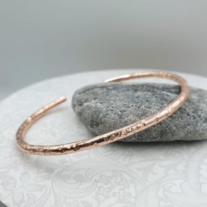 Puede incluir: Un brazalete de cobre martillado con un diseño abierto. El brazalete tiene una superficie texturizada y se muestra sobre una piedra gris. El brazalete es una pieza de joyería simple y elegante.