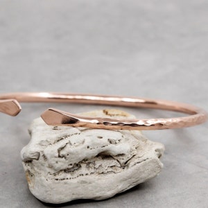 May include: A rose gold cuff bracelet with a hammered texture. The open-ended design features a geometric, angled detail on one end. The bracelet is displayed on a white, textured surface against a grey background.