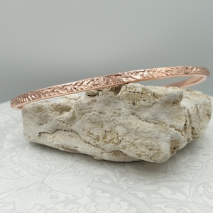 May include: A rose gold bangle with a textured surface, displayed on a light-coloured stone. The bracelet features a hammered or etched design. The overall aesthetic is elegant and minimalist, suitable for everyday wear or special occasions. The bangle's diameter is approximately 6.5 cm.