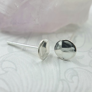 May include: A pair of silver stud earrings with a hammered finish. Each earring features a post and a rounded, uneven design. The background is a patterned white surface with a soft purple hue.