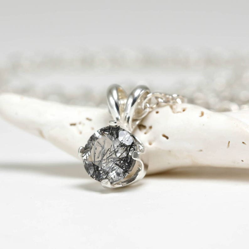 May include: A silver pendant necklace with a round, clear quartz stone with black inclusions. The stone is set in a silver bezel setting.