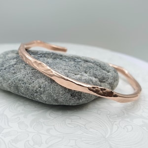 Puede incluir: Un brazalete de puño de oro rosa martillado. El brazalete está hecho de una sola pieza de metal que ha sido martillada para crear una superficie texturizada. El brazalete está abierto y se puede ajustar para que se ajuste a la mayoría de las muñecas.
