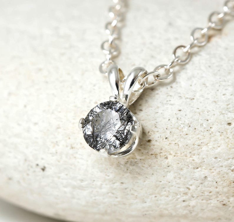 May include: A silver chain necklace with a round, clear quartz pendant with black inclusions. The pendant is set in a silver bezel.
