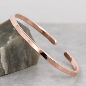 May include: A simple, hammered rose gold bangle bracelet. The bracelet is made from a single piece of metal and has a smooth, polished finish.
