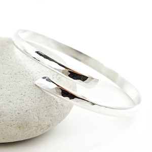 May include: A silver hammered bangle bracelet with a unique open design. The bracelet is made from a single piece of metal that is bent into a circle and then hammered to create a textured surface.