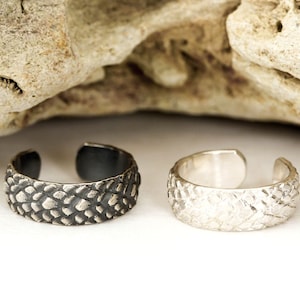 May include: Two adjustable rings with a textured, scaly design. One ring is silver and the other is black.