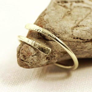 May include: A gold-coloured bypass ring featuring a thin, open-ended band with a textured, hammered finish on the outer surface. The ring is constructed with two overlapping ends that do not meet, creating a continuous spiral-like appearance.