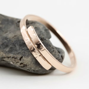 May include: A rose gold bangle bracelet. One side has a hammered texture, the other is smooth. The open design wraps around the wrist. The jewellery is displayed on a grey stone. The bracelet's diameter is approximately 6 cm.
