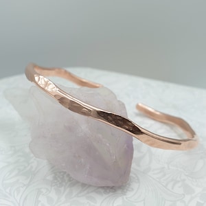 Peut inclure: Un bracelet manchette en cuivre martelé. Le bracelet ouvert a une surface texturée et une forme légèrement irrégulière. La couleur rose-doré chaude du métal contraste avec le fond clair. Le bracelet est présenté sur un cristal violet clair.