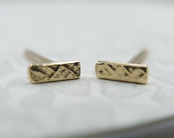 May include: A pair of gold-colored bar stud earrings. Each earring has a textured, rectangular design. The earrings are set against a soft, neutral background, highlighting their simple elegance and minimalist style.