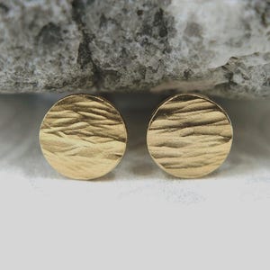 May include: A pair of circular gold stud earrings featuring a textured, rippled surface pattern. The earrings have a polished metallic finish and a flat, round shape that reflects light across the uneven, wavy details on the front face.