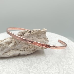 May include: A hammered copper cuff bracelet. The open-ended bracelet has a textured surface and a warm, reddish-brown colour. The bracelet is resting on a piece of light-coloured, textured stone.