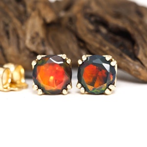 May include: A pair of gold stud earrings featuring round, faceted black opal gemstones. The opals display a fiery red and orange iridescence.