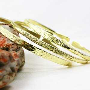 May include: Three gold-coloured bangles with textured surfaces are displayed. The bangles have a hammered metal appearance and are arranged on a white surface, with a portion of a rock visible in the background.