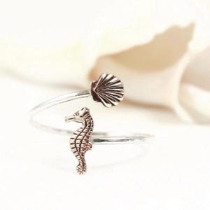 May include: A silver ring featuring a seahorse and seashell motif. The seahorse has textured detailing, and the seashell is fanned. The ring wraps around the finger, offering a distinctive, ocean-inspired accessory.