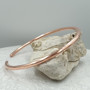 Op de afbeelding: Een koperkleurige, gehamerde metalen manchetarmband. De armband heeft een eenvoudig, open ontwerp met een afgeronde vorm. Het metaal heeft een getextureerd oppervlak en de armband wordt gepresenteerd op een lichtgekleurde steen.