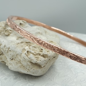 May include: A thin, circular bangle bracelet crafted from rose-coloured metal. The exterior surface of the bangle features a distinct, uneven, and rugged hammered texture that catches the light, contrasting with the smooth inner band.