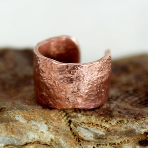 May include: A hammered copper ring with a textured surface. The ring is wide and has a rounded shape.