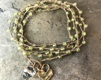 Puede incluir: Pulsera envolvente de múltiples hilos con cuentas verde claro y un dije de corazón dorado. La pulsera está hecha con un cordón de color neutro e incluye un dije de cristal transparente facetado. La pulsera se muestra sobre una superficie de piedra gris.