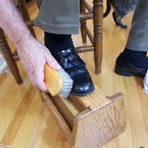 Könnte beinhalten: Eine Person reinigt einen schwarzen Lederschuh mit einer Holzschuhbürste. Der Schuh liegt auf einem hölzernen Schuhputzständer.
