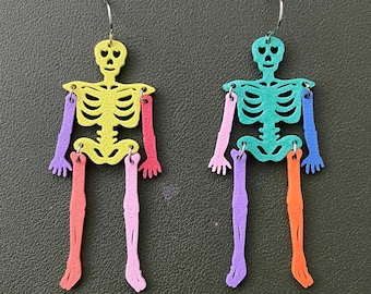 May include: A pair of dangle earrings featuring articulated skeleton figures. The left skeleton has a yellow torso and head, purple arms, and pink legs. The right skeleton has a teal torso and head, pink and blue arms, and purple and orange legs.