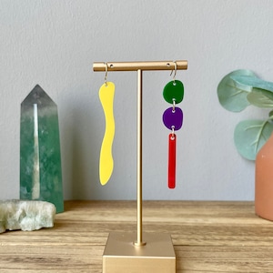 May include: A pair of colorful earrings on a gold stand. One earring is a long, yellow, abstract shape. The other earring has three round shapes: green, purple, and red.