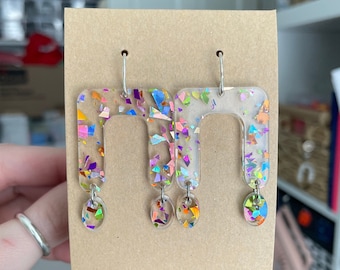 May include: A pair of clear resin earrings with a rainbow confetti design. The earrings are shaped like a rectangle with a rounded top and bottom. Each earring has a small, round dangle charm.