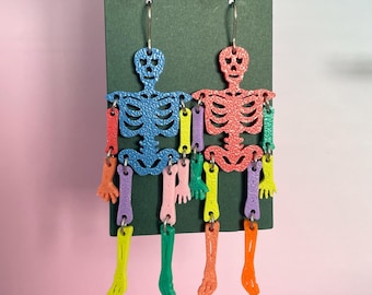 May include: A pair of dangle earrings featuring two articulated skeleton figures. The left skeleton is blue with multicolored limbs, while the right skeleton is coral with multicolored limbs. Each skeleton has a textured surface and silver-tone hooks.