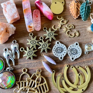 May include: A collection of jewelry-making supplies, including various charms, beads, and pendants. The assortment features gold-toned crescent moons, starbursts, and Hamsa hand charms. Also included are polished stones in shades of pink and orange, and leaf-shaped pendants.